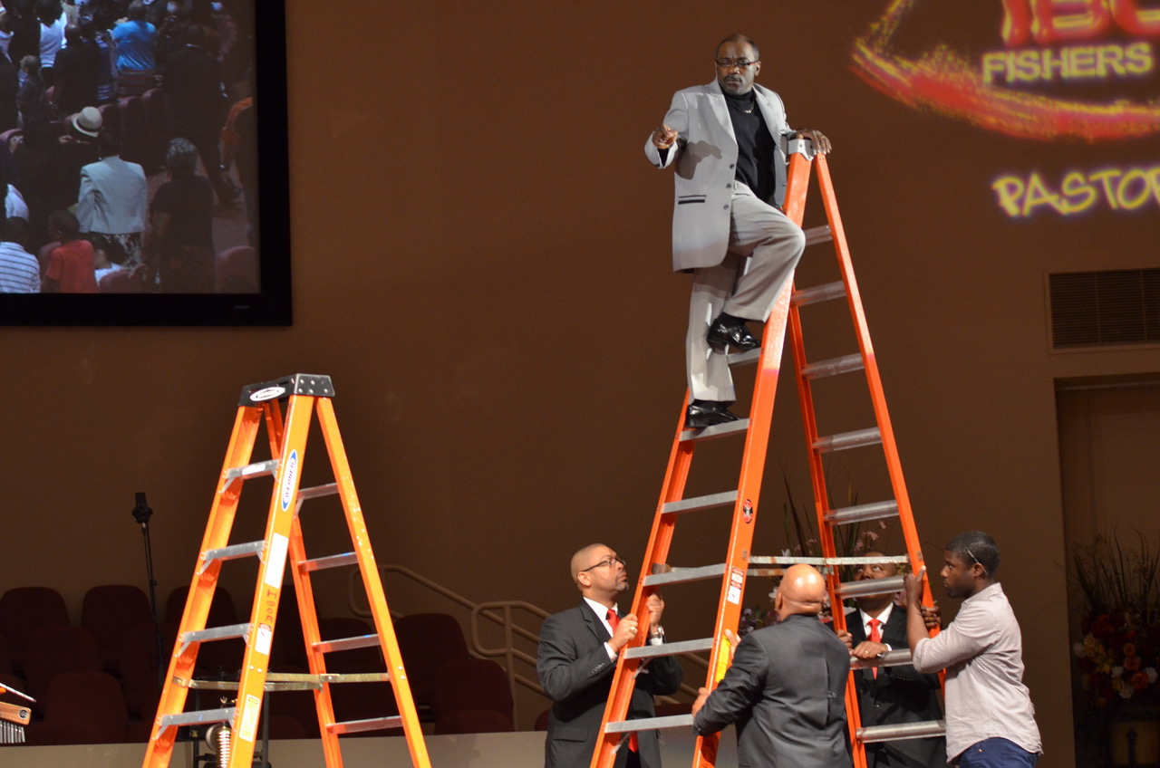 A man in a grey suit standing at the top of a large, orange ladder on a stage. There are 3 men in suits holding the ladder.